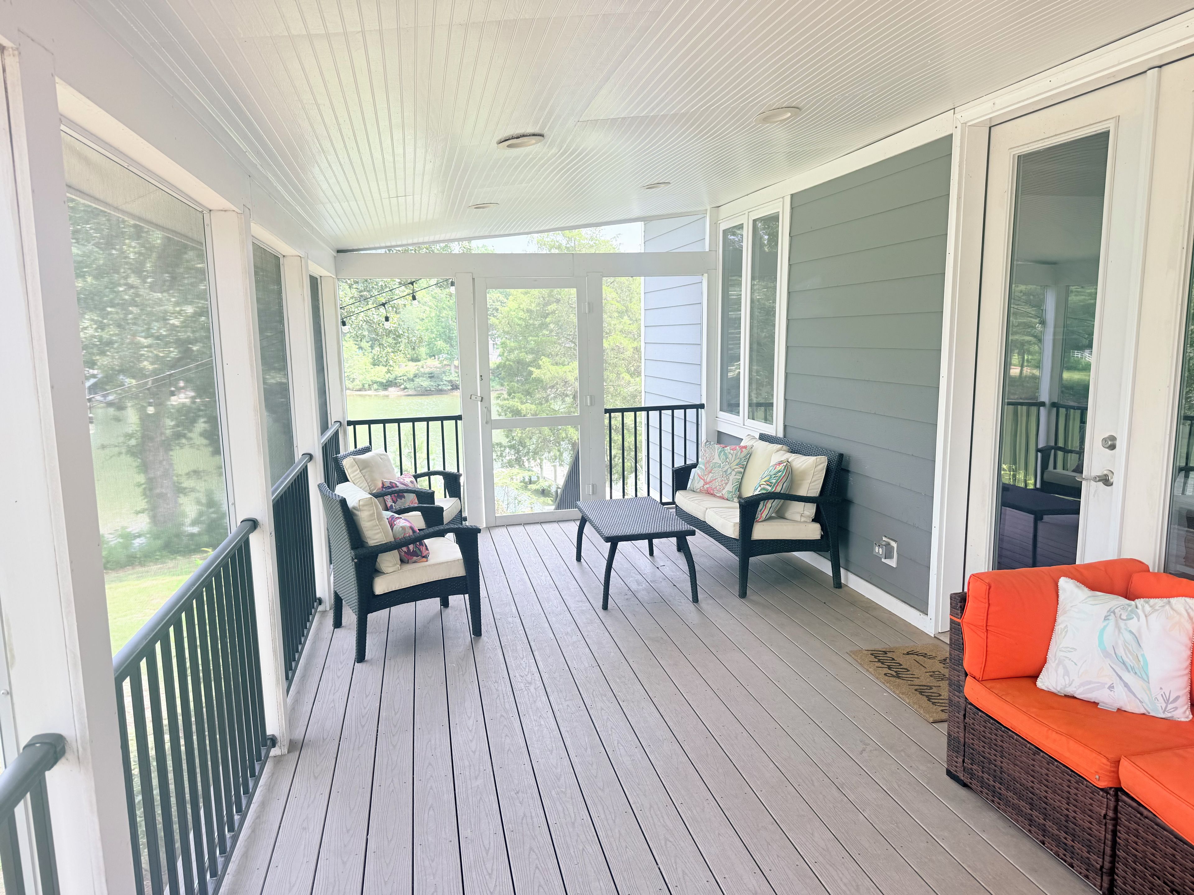 A covered porch with a wooden floor, two wicker chairs with cushions, a wicker sofa, and a red cushioned bench.