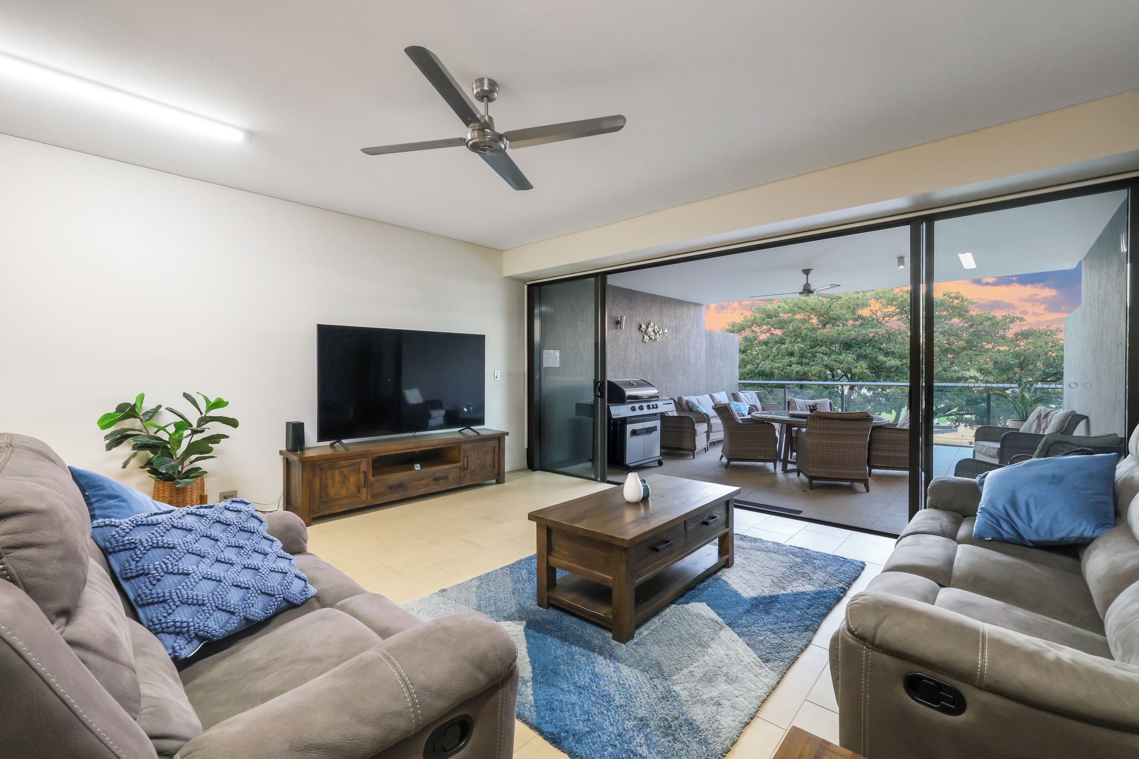 A modern living room with a flat-screen TV, a ceiling fan, and a view of an outdoor patio with furniture.