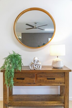 A wooden console with a round mirror, a ceiling fan, a potted plant, rolled towels, and a lamp. - Lagoon Glow Waterfront Sunsets and Poolside Days (Darwin)