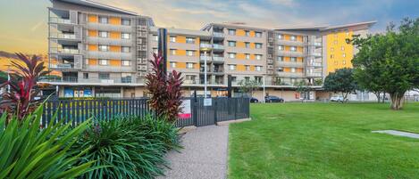 A multi-story residential building with balconies, a paved pathway, and a grassy area.