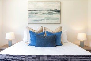 A bed with a white duvet, two side tables with lamps, and a painting of a seascape above the bed. - Lagoon Glow Waterfront Sunsets and Poolside Days (Darwin)