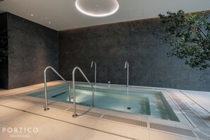 Uma área de piscina interna moderna com uma parede de pedra escura, piscina cristalina e escada metálica.