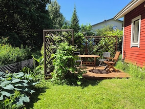 En træterrasse med et bord og stole, omgivet af frodig vegetation og en rød bygning.