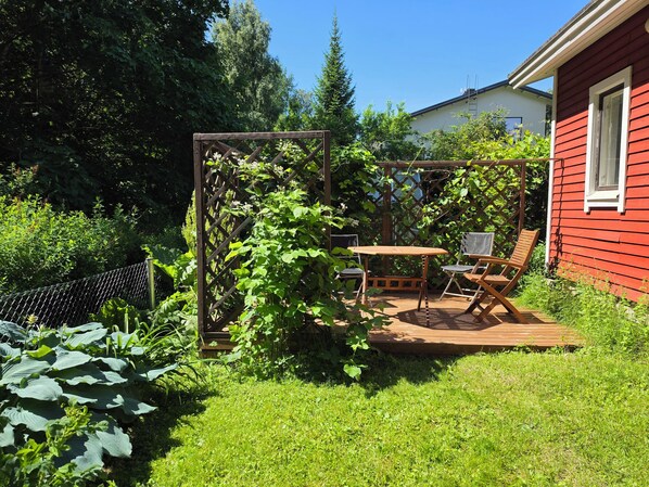 A wooden deck with a table and chairs, surrounded by lush greenery and a red building.