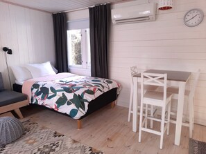 A room with a bed, a small table, a chair, an air conditioning unit, and a clock on the wall.