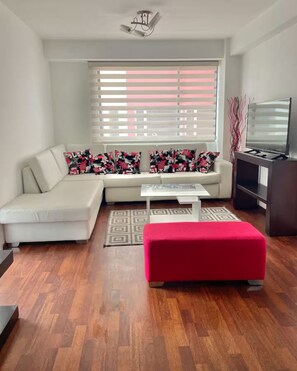 Ein Wohnzimmer mit einem weißen Sofa, einem roten Pouf, einem Couchtisch und einem TV-Schrank.