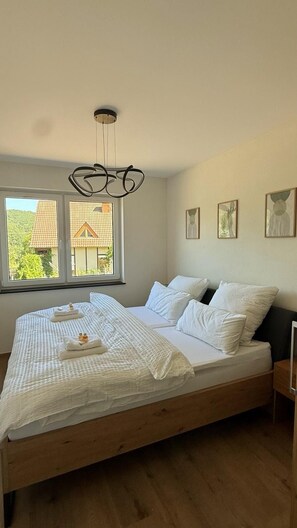 A bedroom with a wooden bed, white bedding, a hanging light fixture, and a window with a view of houses. - Ferienhaus Grünblick (Mauel)