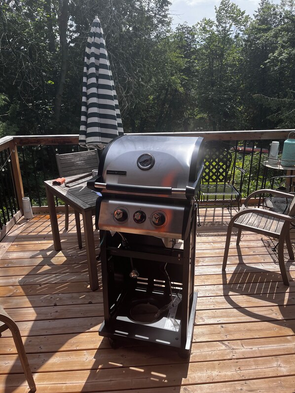 A barbecue grill on an outdoor deck with a wooden table and chairs, surrounded by trees.
