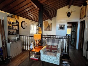 A cozy living room with a wooden ceiling, a fireplace, a dining table with a lamp, and a chair with a Union Jack pillow.