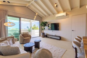 A modern living room with a large window, a flat-screen TV mounted on the wall, and a ceiling fan. - Solin at The Reef Superior - Ocean View Penthouse (Willemstad)