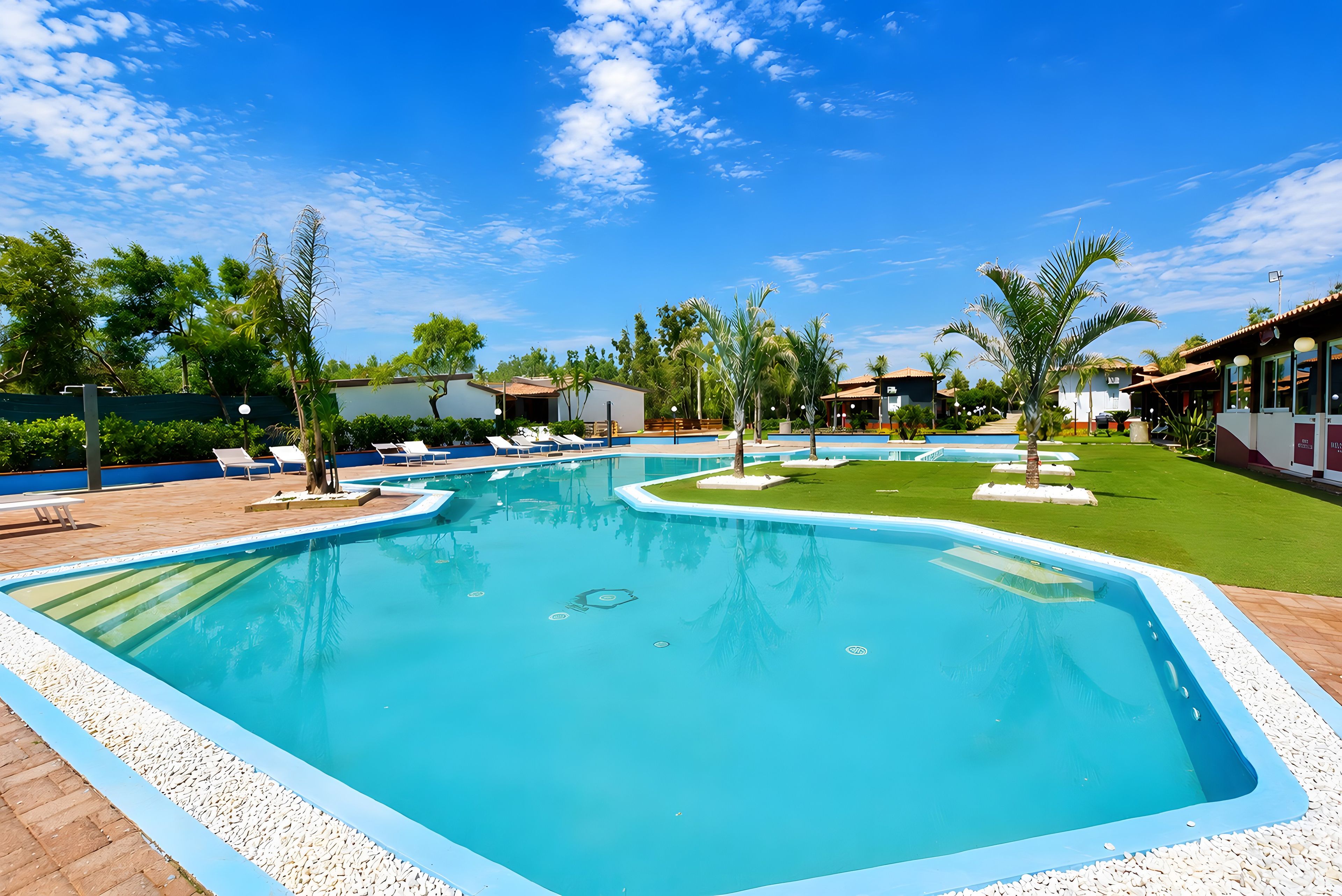 Ein Swimmingpool mit Liegestühlen und Palmen, umgeben von Gebäuden und wolkenlosem Himmel.