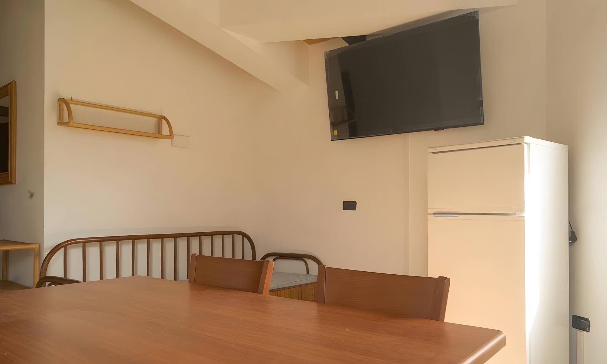 A dining area with a wooden table and chairs, a television mounted on the wall, and a refrigerator.