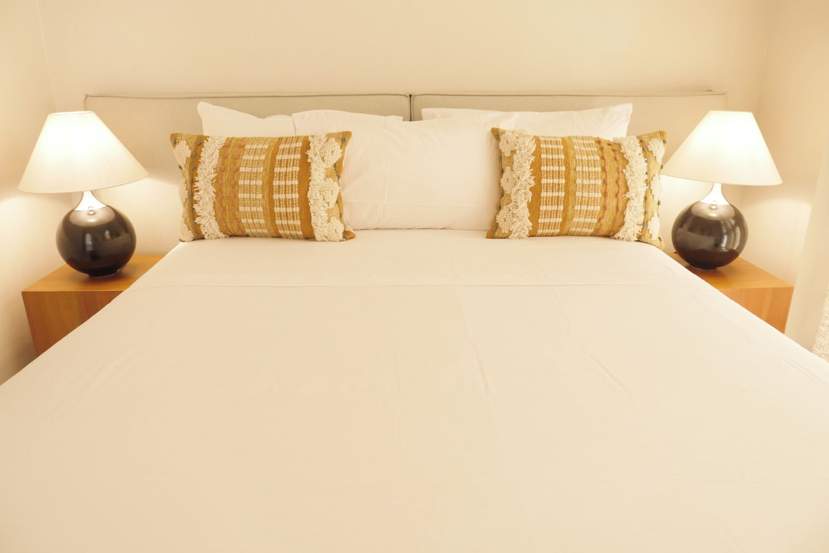 Una cama bien hecha con sábanas blancas, dos almohadones decorativos y dos lámparas de noche sobre mesitas de madera.