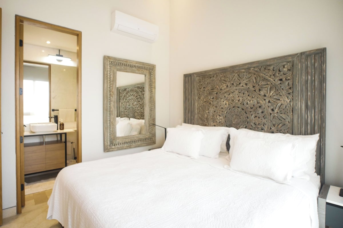 Habitación de hotel con una cama grande, un espejo, cabecera de madera y un baño visible a través de una puerta abierta.