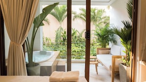 A hotel room with a bed, a small table, a sofa, a hot tub, and a view of a garden with various plants. - Casa Pedregal 301 (Cartagena de Indias)