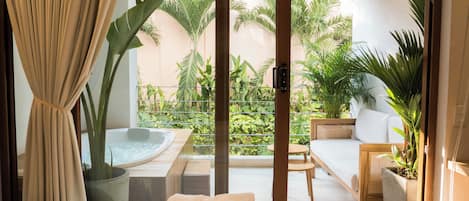 A hotel room with a bed, a small table, a sofa, a hot tub, and a view of a garden with various plants.