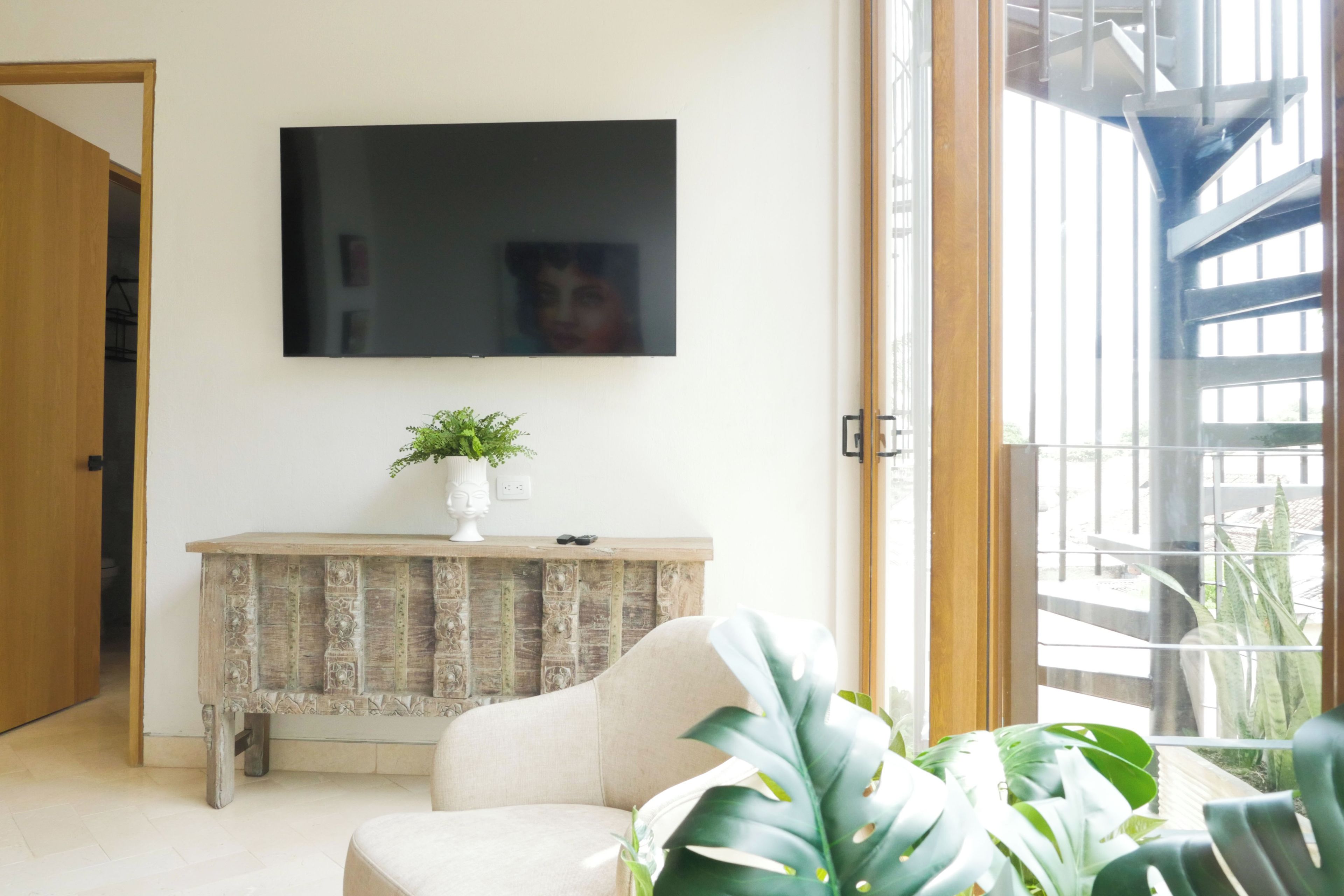 A living room with a flat-screen TV mounted on the wall, a wooden console table with a plant, and a large window offering a view of the outdoors.