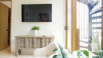 A living room with a flat-screen TV mounted on the wall, a wooden console table with a plant, and a large window offering a view of the outdoors.