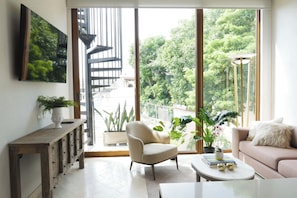 A modern living room with a sofa, armchair, coffee table, and large windows offering a view of greenery and a spiral staircase. - Casa Pedregal (Cartagena de Indias)
