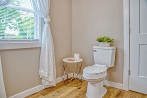A small bathroom with a toilet, a small table with a roll of toilet paper, a potted plant on the toilet tank, and a window with curtains. - Cascade Haven - Near Cummins Falls & Cookeville (Cookeville)