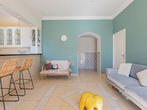 A living room with a sofa, a dining area with chairs, a kitchen with cabinets, and a yellow elephant toy on the floor. - 3 chambres & terrasse - Aux portes de Monaco (Beausoleil)