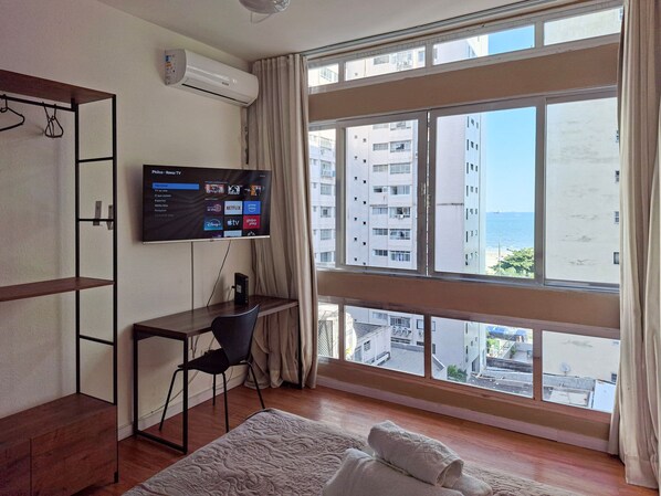 A hotel room with a large window, a flat-screen TV mounted on the wall, a desk with a chair, and a bed with a white duvet.