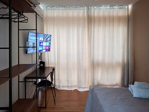 Habitación de hotel con cama, escritorio, computadora, televisor y ventana con cortinas.