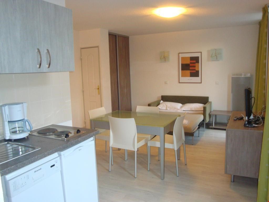 Een compacte hotelkamer met een kitchenette, een bed met kussens, een bureau met een computer en een kleine tafel met stoelen.