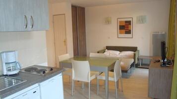 Quarto de hotel compacto com cozinha compacta, uma cama com travesseiros, uma mesa com computador e uma pequena mesa com cadeiras.