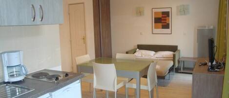 Une chambre d’hôtel compacte comprenant un coin cuisine, un lit avec des oreillers, un bureau avec un ordinateur et une petite table avec des chaises.
