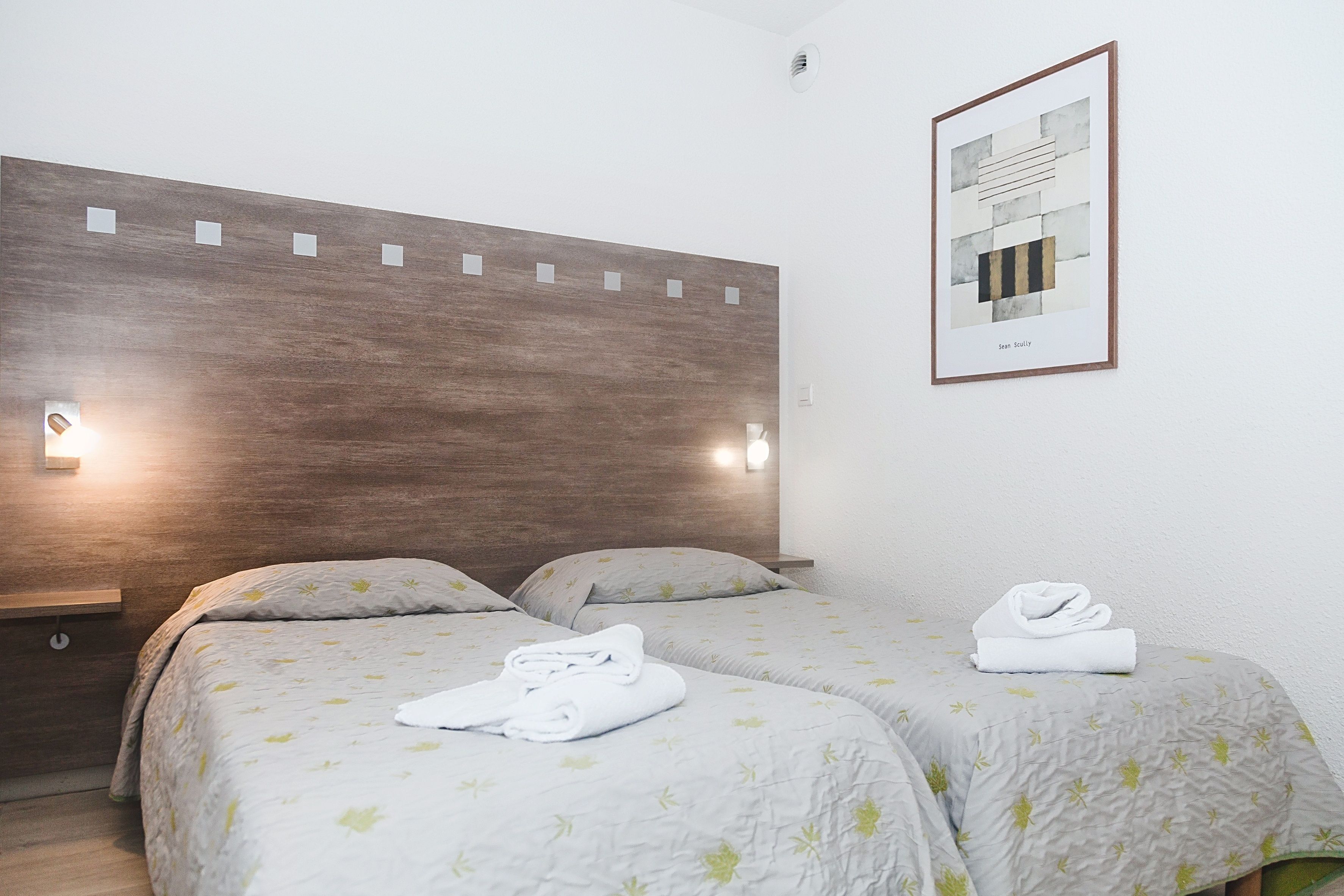 Habitación de hotel con dos camas, cabecera de madera, luces fijadas a la pared y una obra de arte enmarcada.