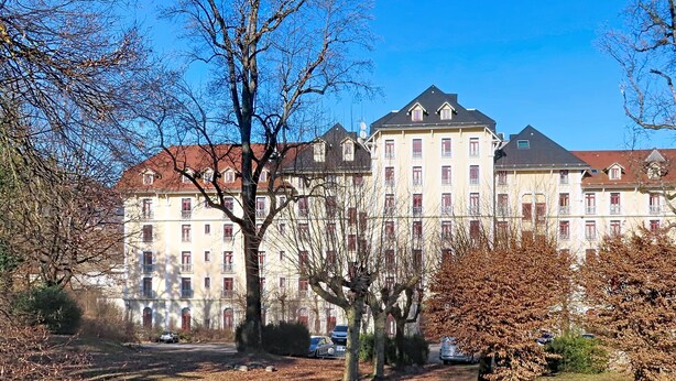 Ein großes Gebäude mit zahlreichen Fenstern und Balkonen, umgeben von Bäumen und einer Rasenfläche.