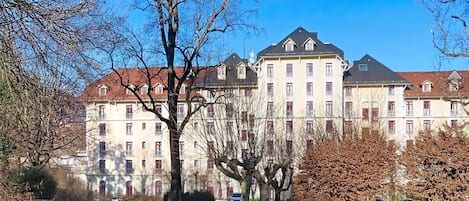 Un grand bâtiment doté de nombreuses fenêtres et de balcons, entouré d’arbres et d’une zone herbeuse.