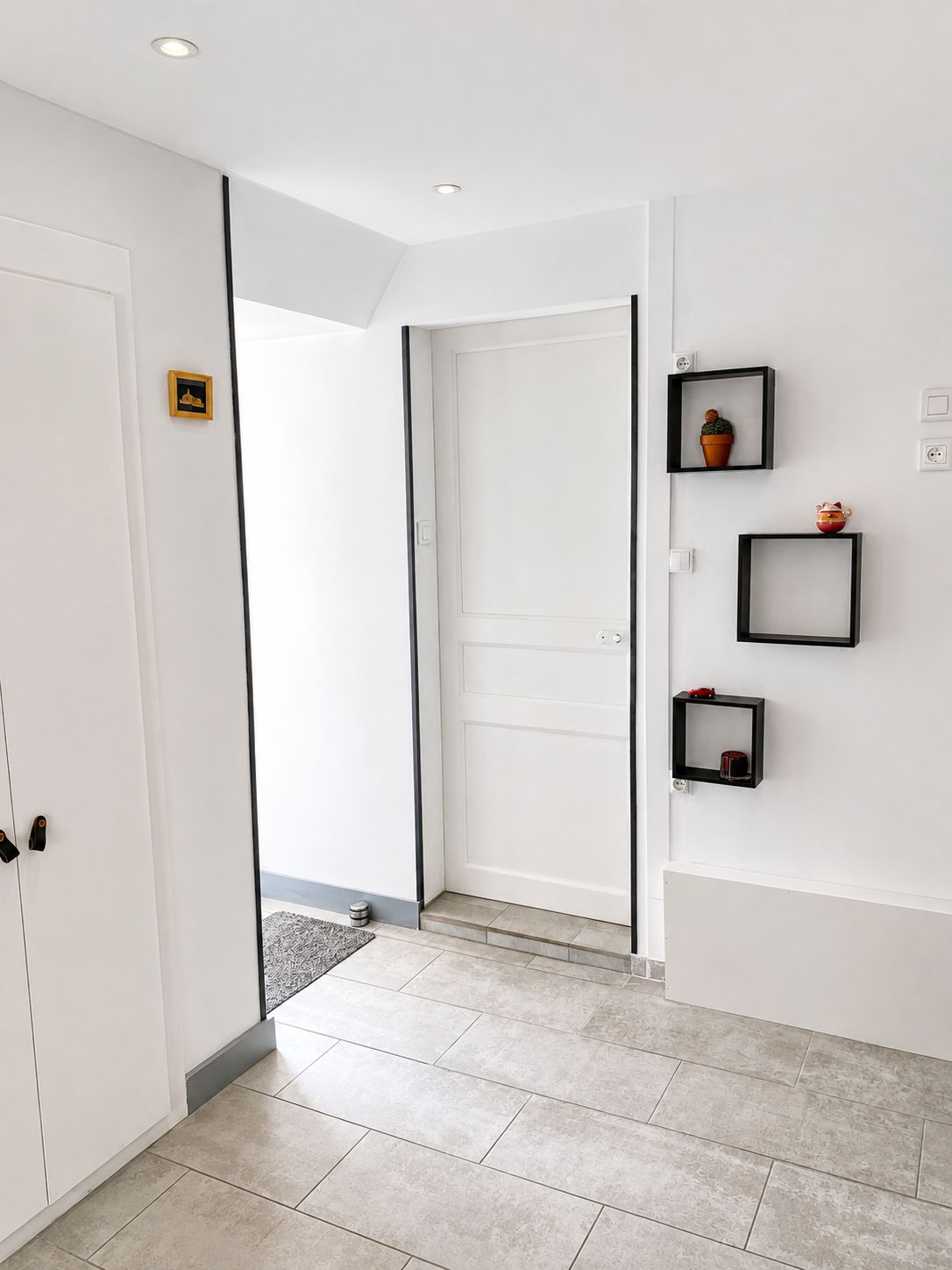 A hallway with a white door, a small framed picture, and three black-framed shelves with decorative items.