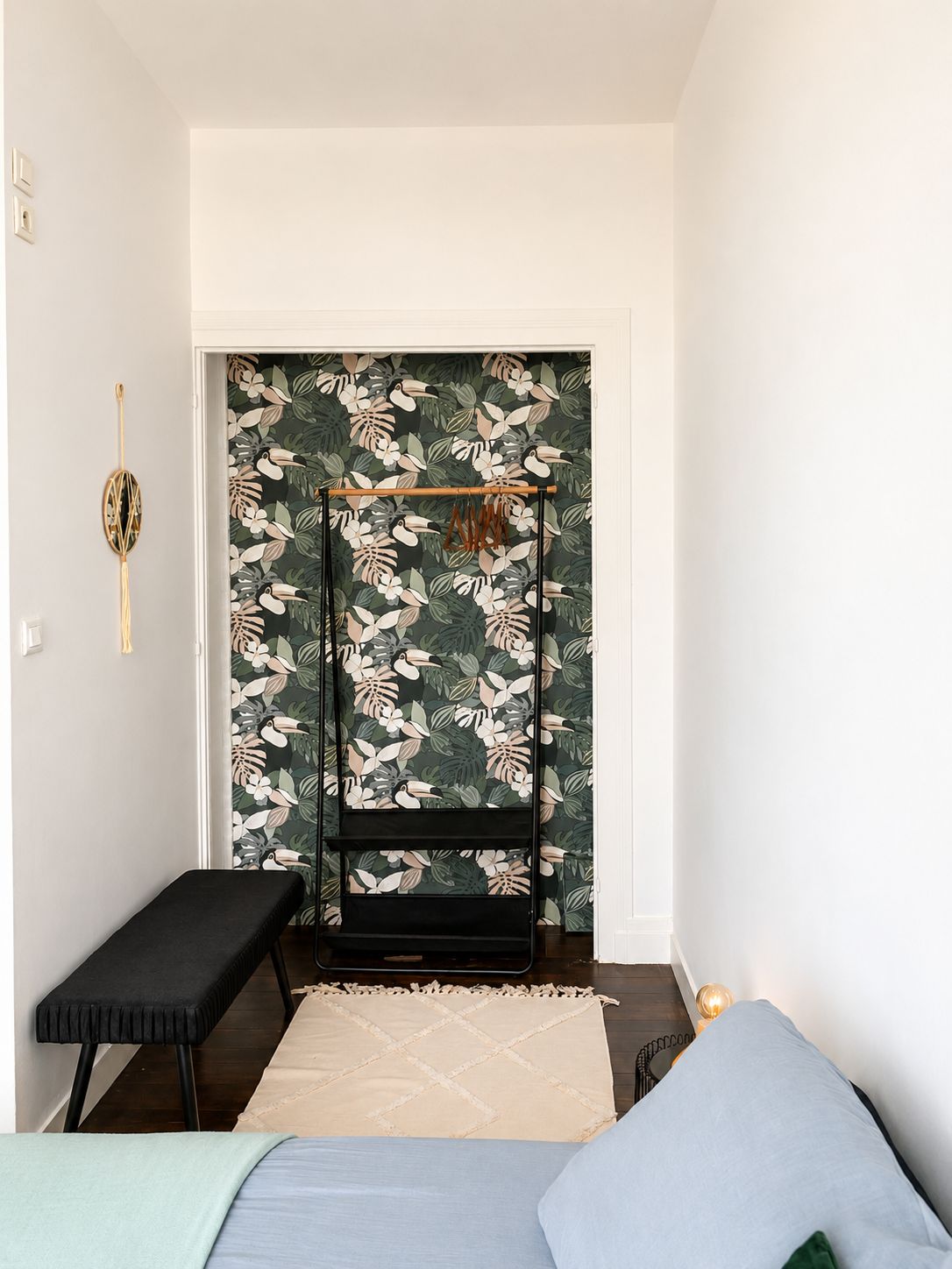 Une pièce comprenant un lit, un banc, une étagère fixée au mur et un papier peint à motifs.