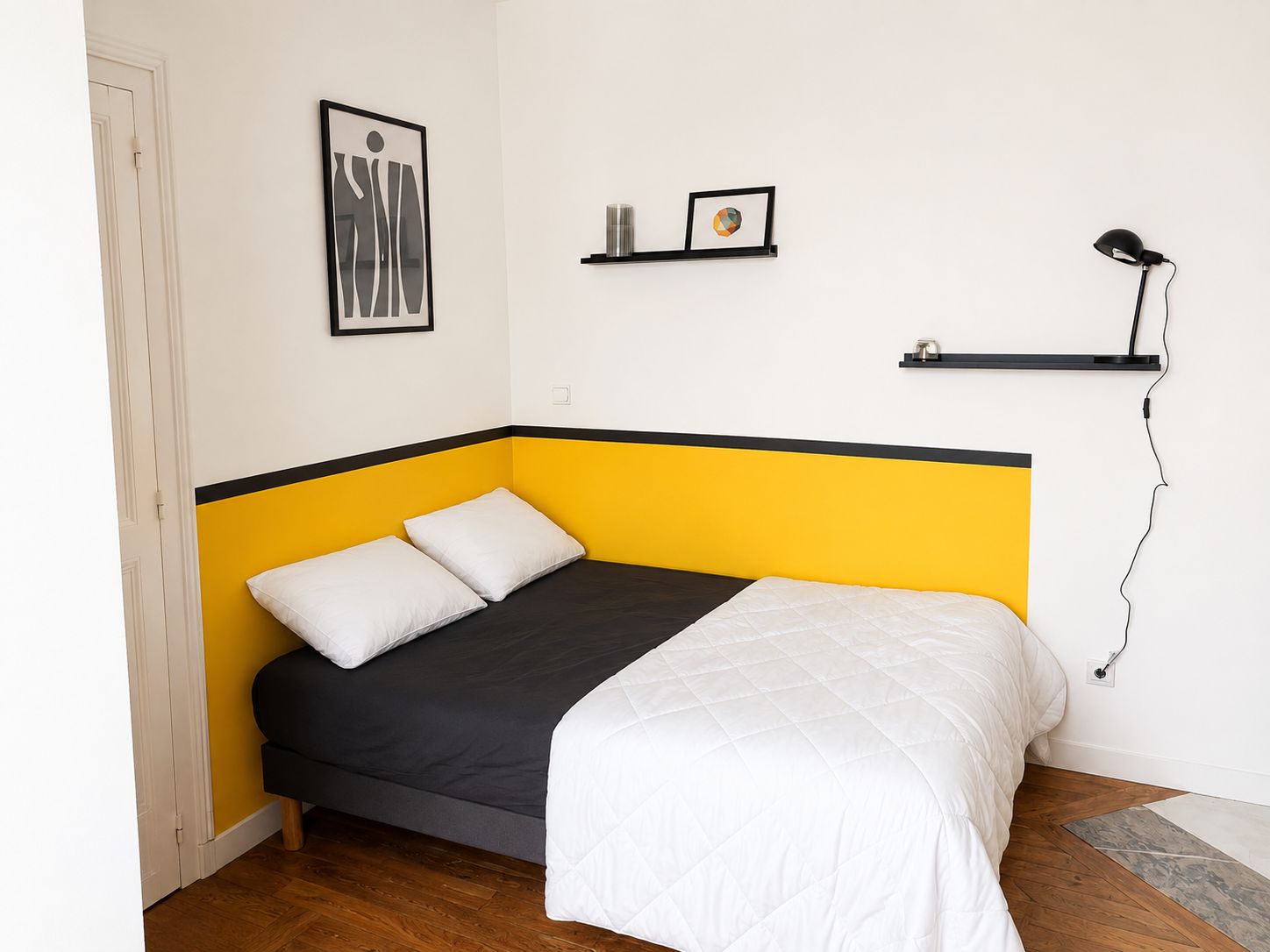 Ein kleines Zimmer mit einem Einzelbett, einer Wand in Gelb und Schwarz sowie einem Bettüberwurf in Weiß und einer schwarzen Schreibtischlampe.