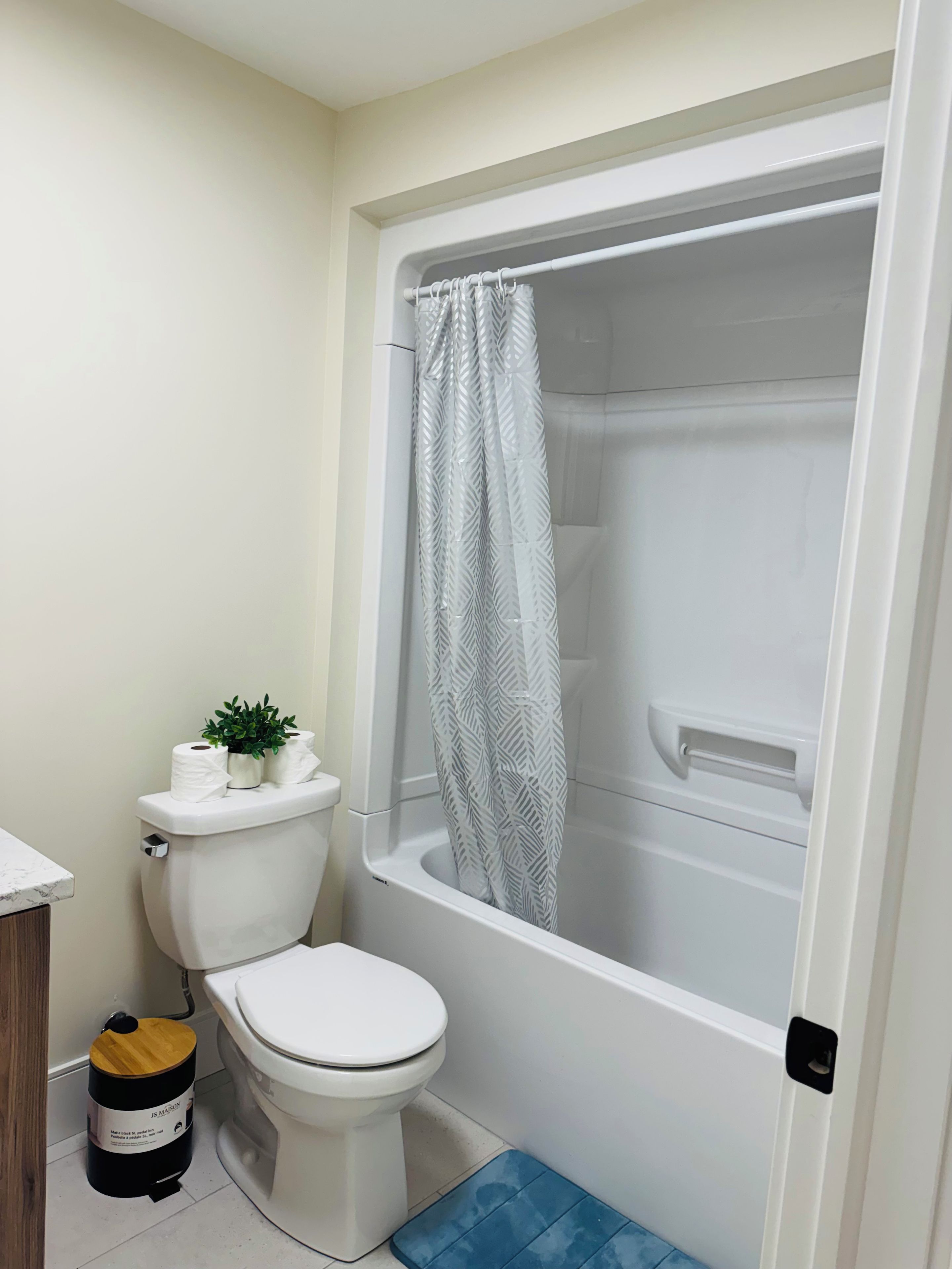 A bathroom with a toilet, a shower with a patterned curtain, and a small plant on the toilet tank.