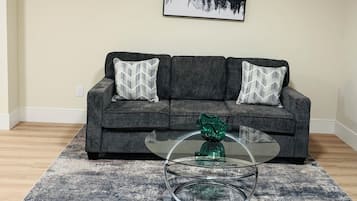 A living room with a grey sofa, two patterned throw pillows, a glass coffee table with a green vase, and a framed landscape painting on the wall.