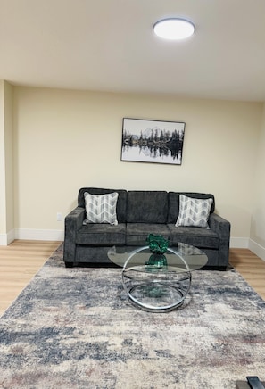 A living room with a grey sofa, two patterned throw pillows, a glass coffee table with a green vase, and a framed landscape painting on the wall.
