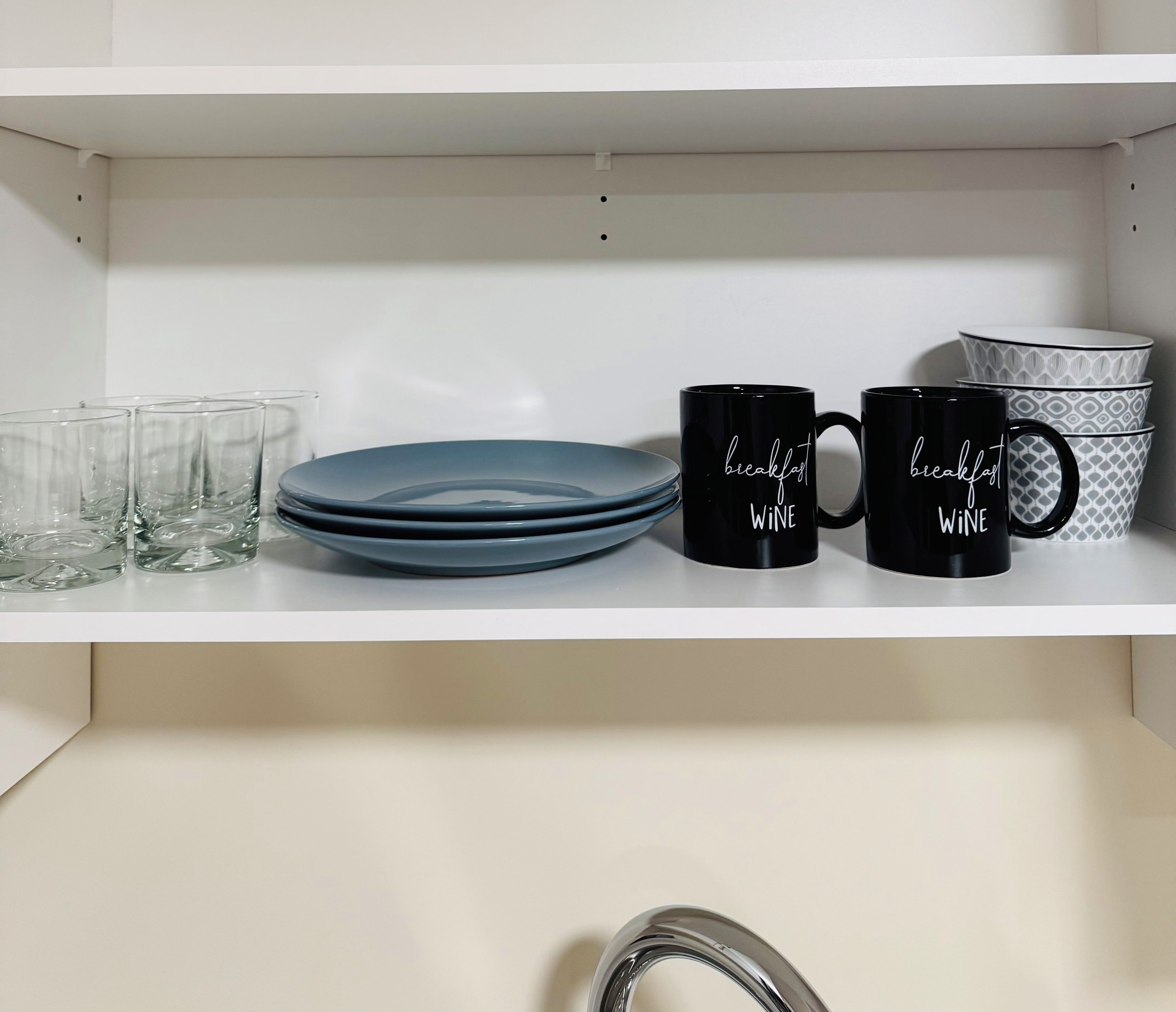 Een keukenkastje met gestapelde borden, twee zwarte mokken met de tekst 'breakfast wine' en een beker met een patroon.