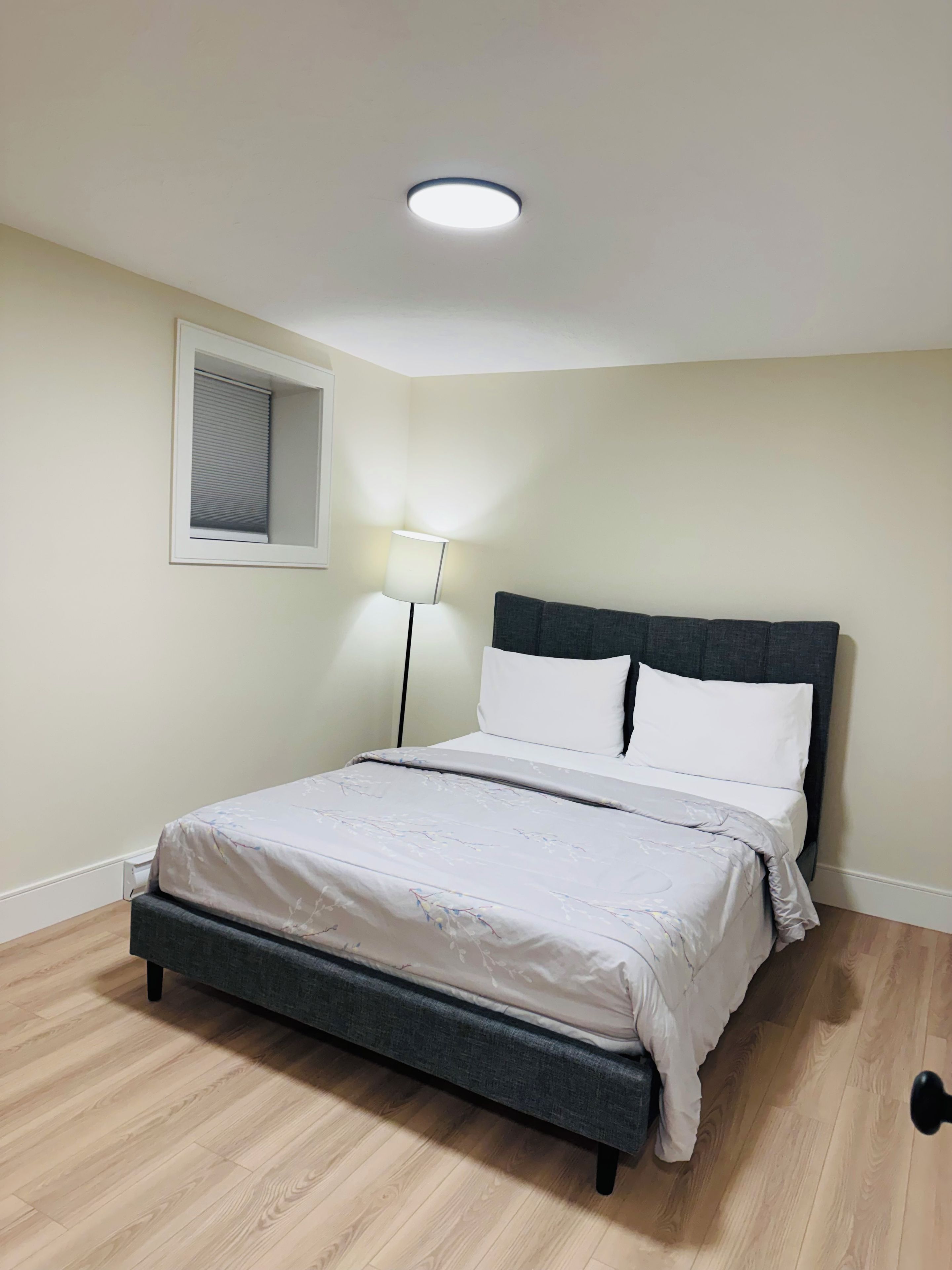 A bedroom with a bed, a bedside lamp, a window with blinds, and a ceiling light.
