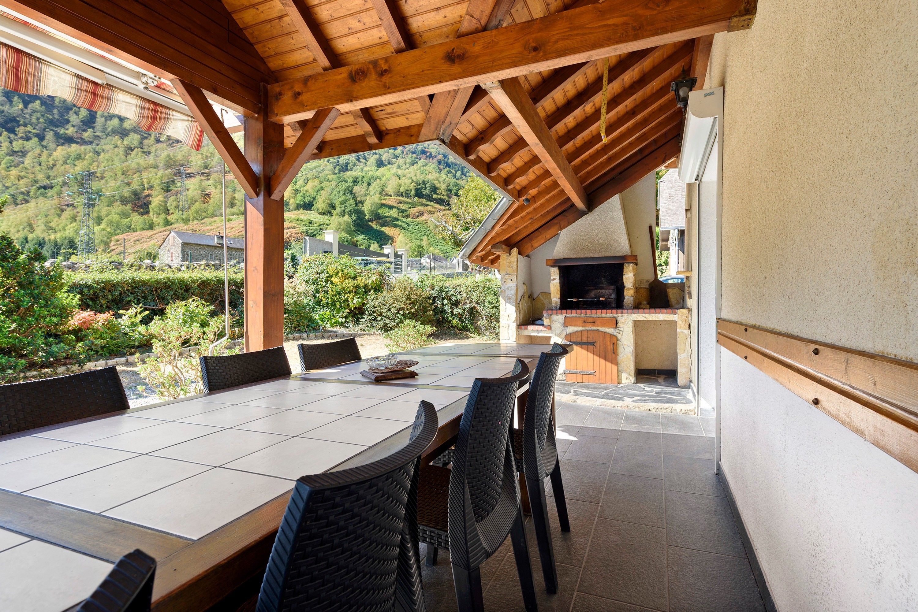 An outdoor dining area with a long table and chairs, a stone fireplace, and a view of a hilly landscape.