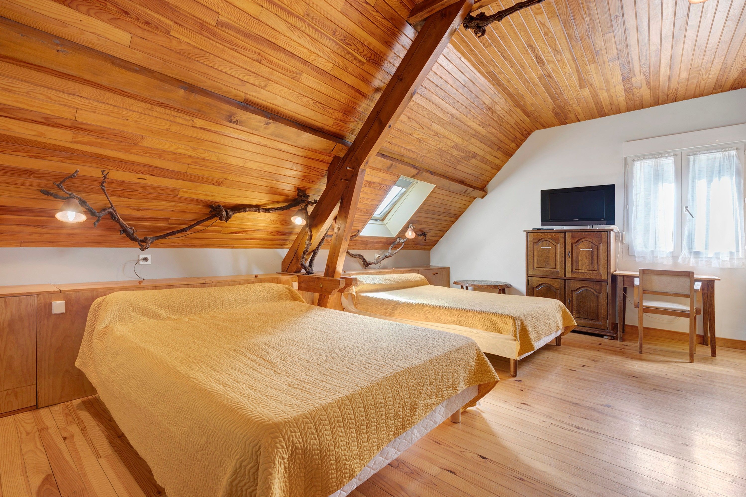 Une pièce avec un plancher en bois, deux lits avec une literie jaune, une armoire en bois, un téléviseur posé sur un meuble et une fenêtre avec des rideaux.