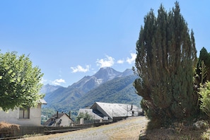 A rural setting with a mountain backdrop, a small building with a sloped roof, and a tall coniferous tree.