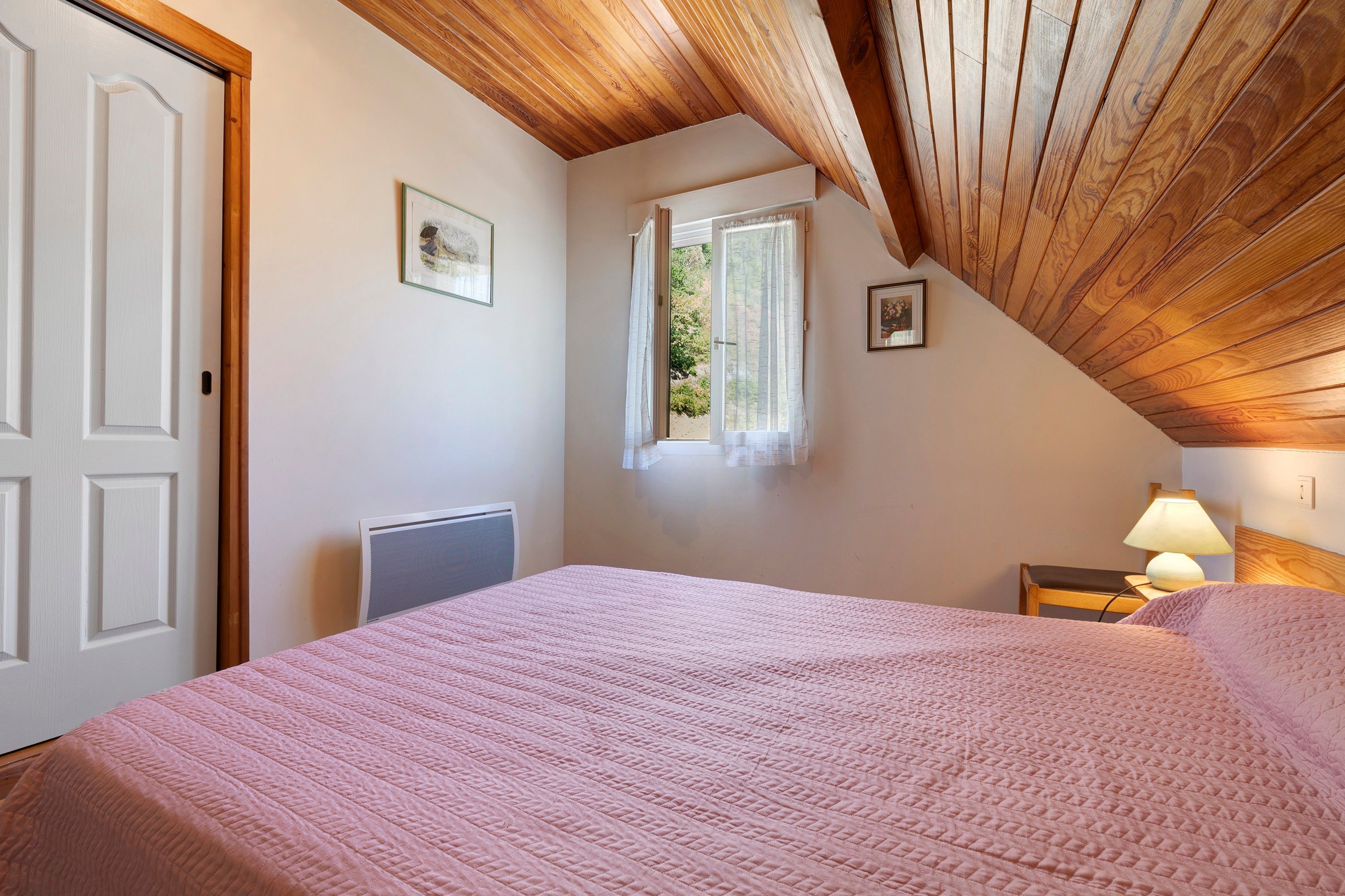 Una camera da letto con soffitto spiovente, un letto con copripiumino rosa, una finestra con vista e una lampada da comodino.