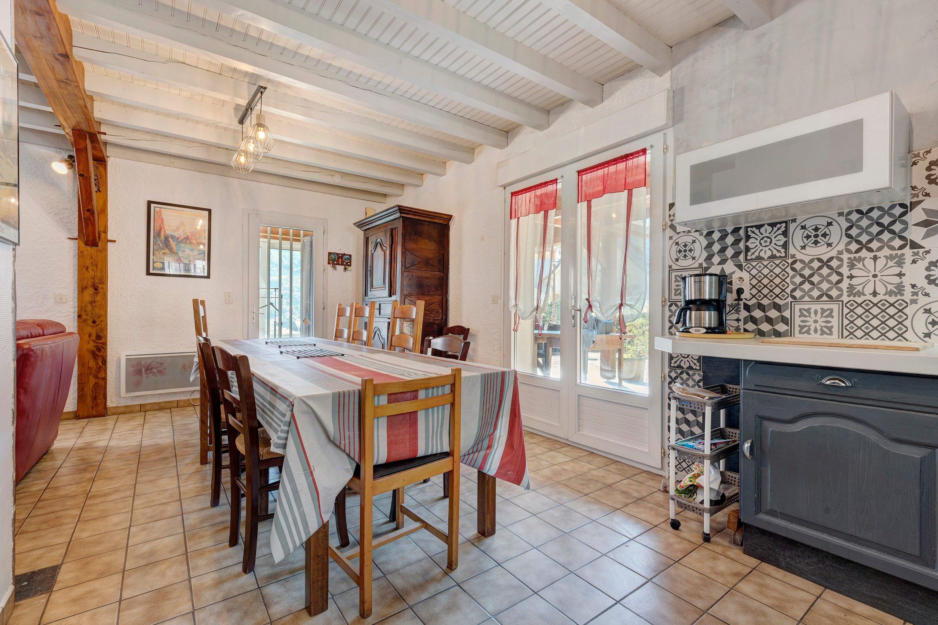 A dining area with a wooden table and chairs, a kitchen with patterned tiles, and a view through the windows.
