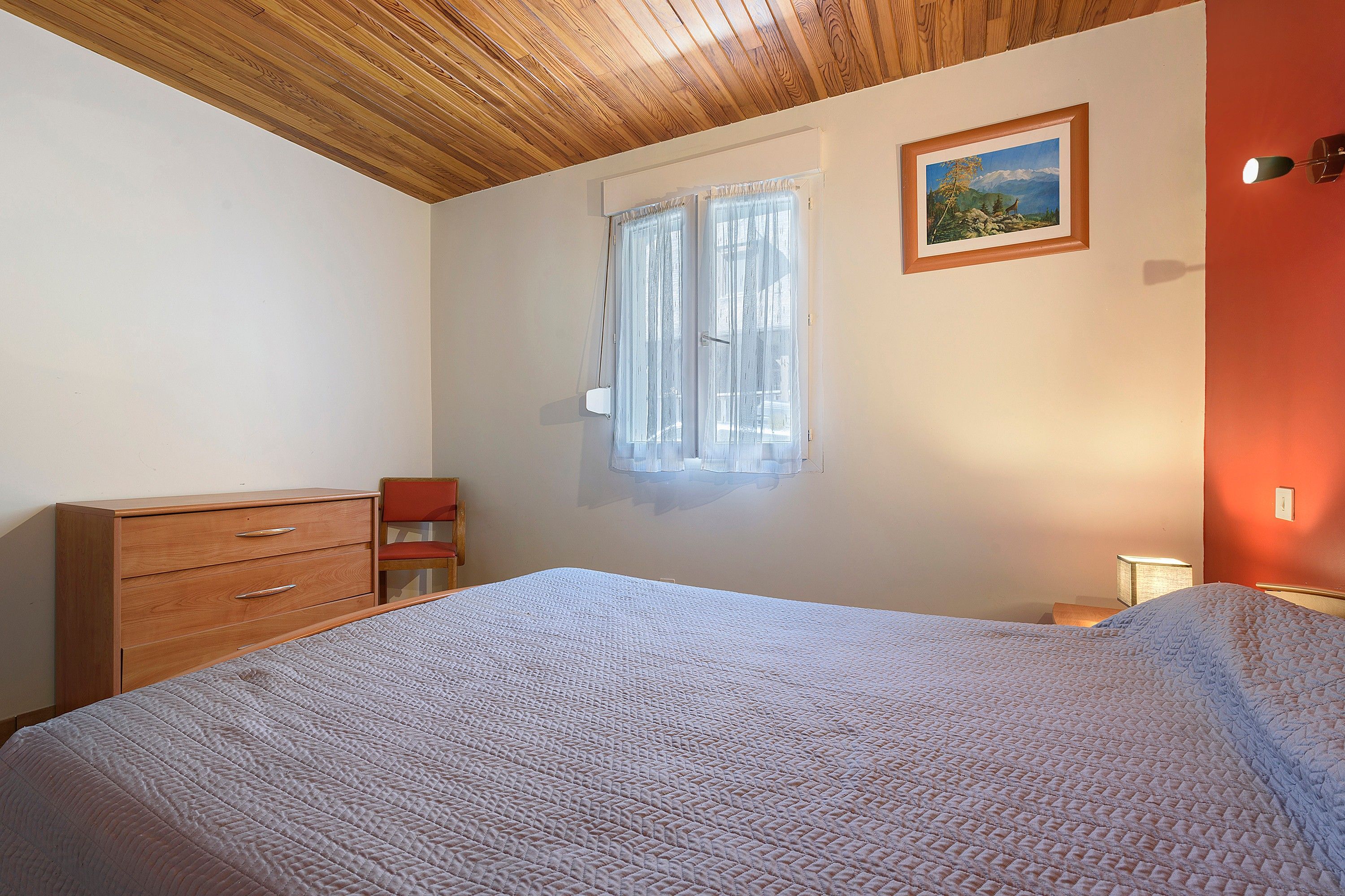 Un dormitorio con techo de madera, una cama con colcha estampada, un aparador, una silla, un ventanal con cortinas traslúcidas y un cuadro enmarcado en la pared.