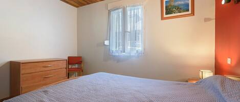 Un dormitorio con techo de madera, una cama con colcha estampada, un aparador, una silla, un ventanal con cortinas traslúcidas y un cuadro enmarcado en la pared.