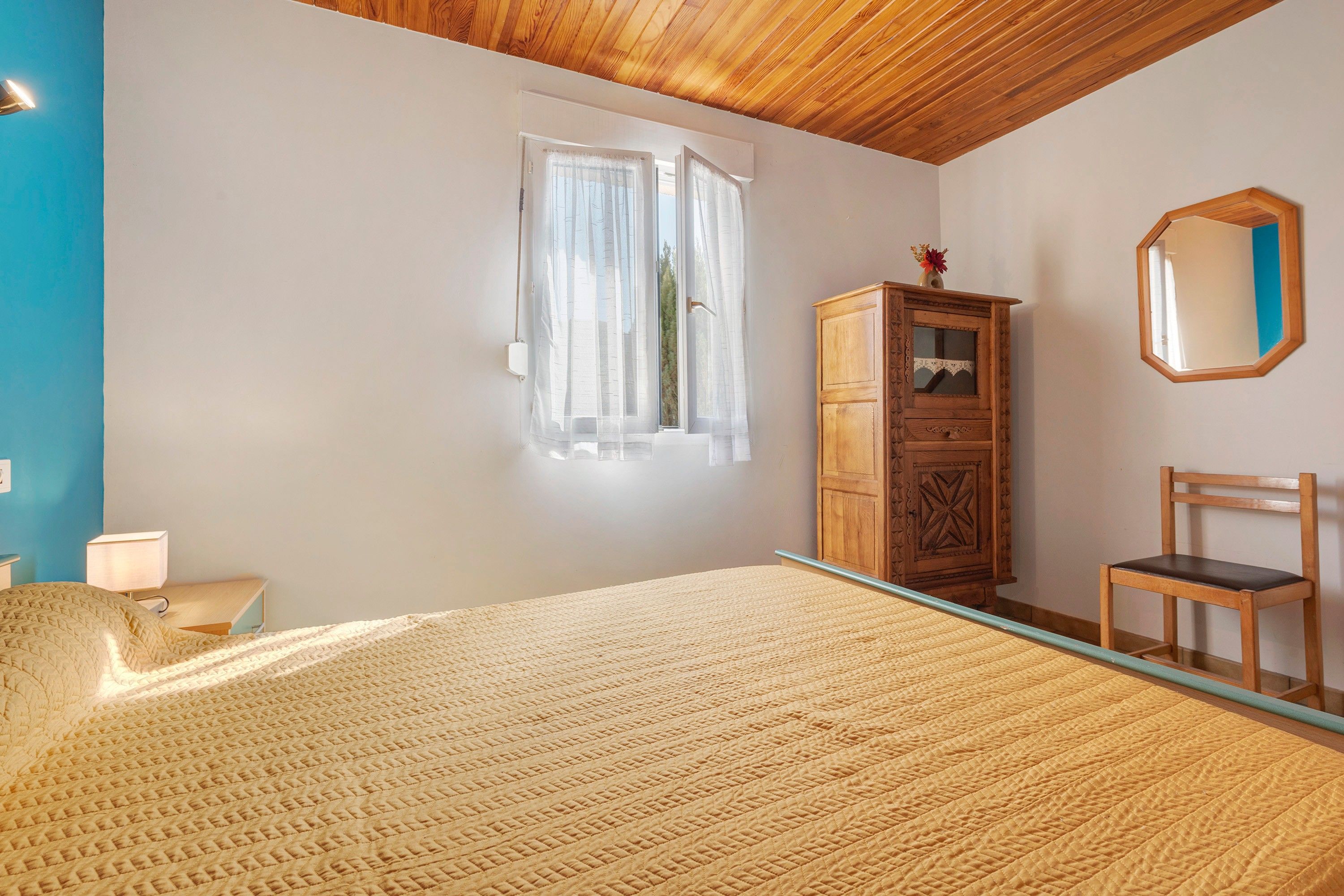 Une chambre à coucher avec un lit, une armoire en bois, une chaise, un miroir et une fenêtre avec des rideaux.