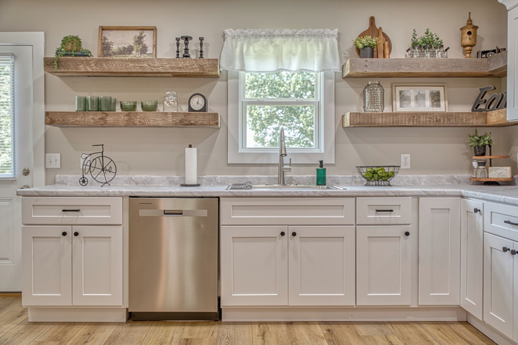 A kitchen with white cabinets, a stainless steel dishwasher, a window with a view, and shelves with various items.